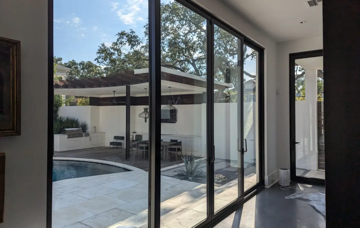 View from inside a modern home with large glass doors, showcasing a pool area and outdoor kitchen surrounded by greenery